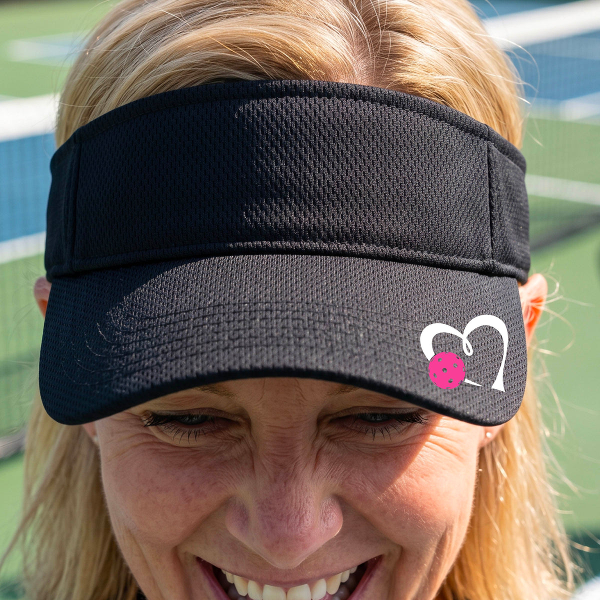 A woman wearing a black athletic visor with a white heart outline and a pink heart and pickleball graphic on the brim, photographed outdoors on what appears to be a pickleball court.