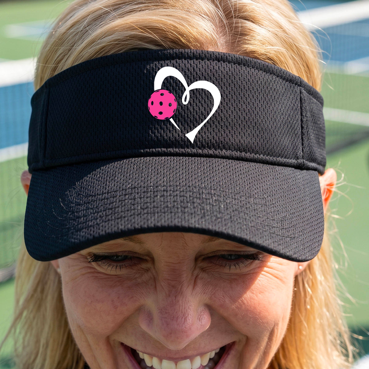 A woman wearing a black athletic visor with a white heart outline and a pink heart and pickleball graphic on the brim, photographed outdoors on what appears to be a pickleball court.
