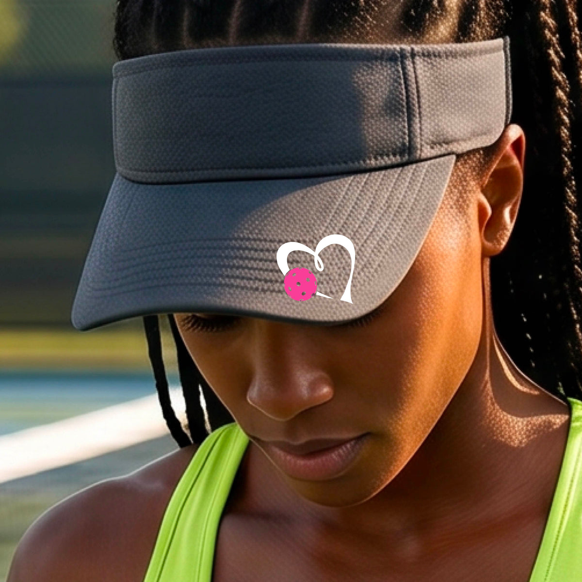 A woman wearing a dark gray athletic visor with a white heart outline and a pink heart and pickleball graphic on the brim, photographed outdoors on what appears to be a pickleball court.