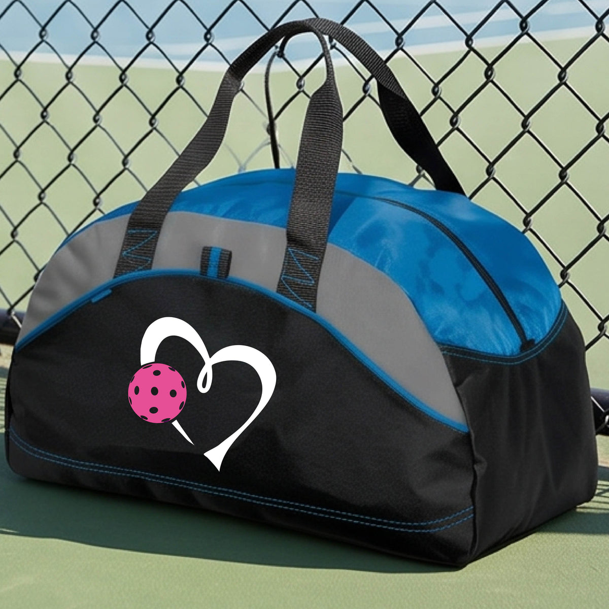 A black and royal blue pickleball duffel bag with gray panels, featuring a white heart outline and a pink pickleball graphic on the front,.