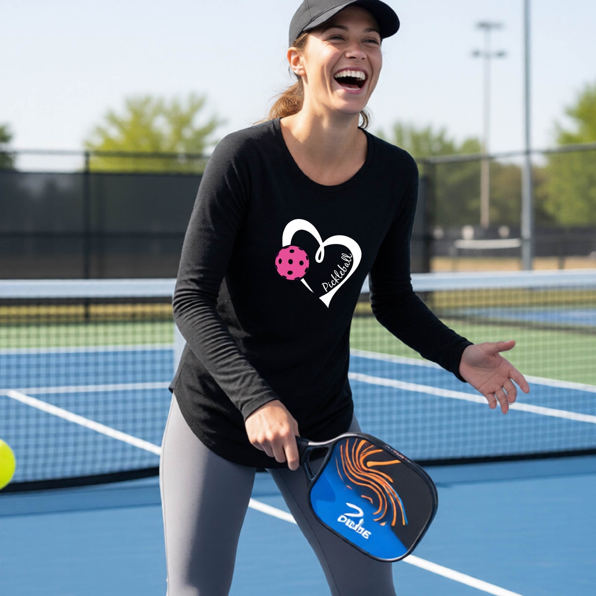 A woman laughing on an outdoor pickleball court wearing a black long-sleeve t-shirt with a white heart graphic and a pink pickleball inside the heart along with the text 'Pickleball' inside the heart.