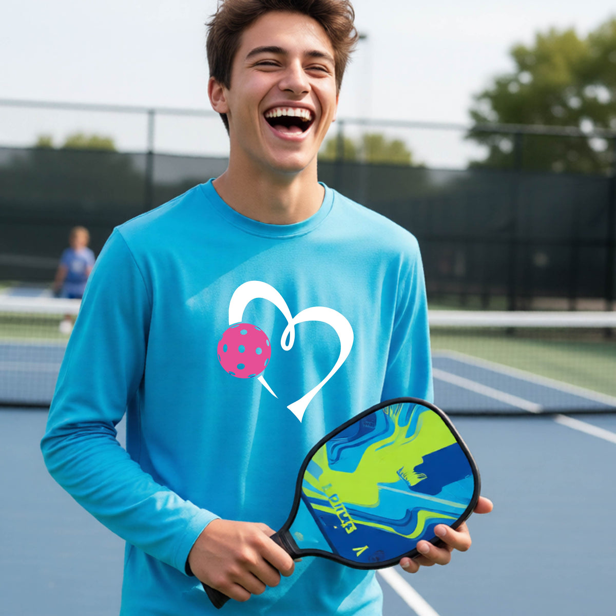 A teenager laughing on an outdoor pickleball court, wearing a bright blue long-sleeve shirt with a white heart and pink pickleball graphic,.