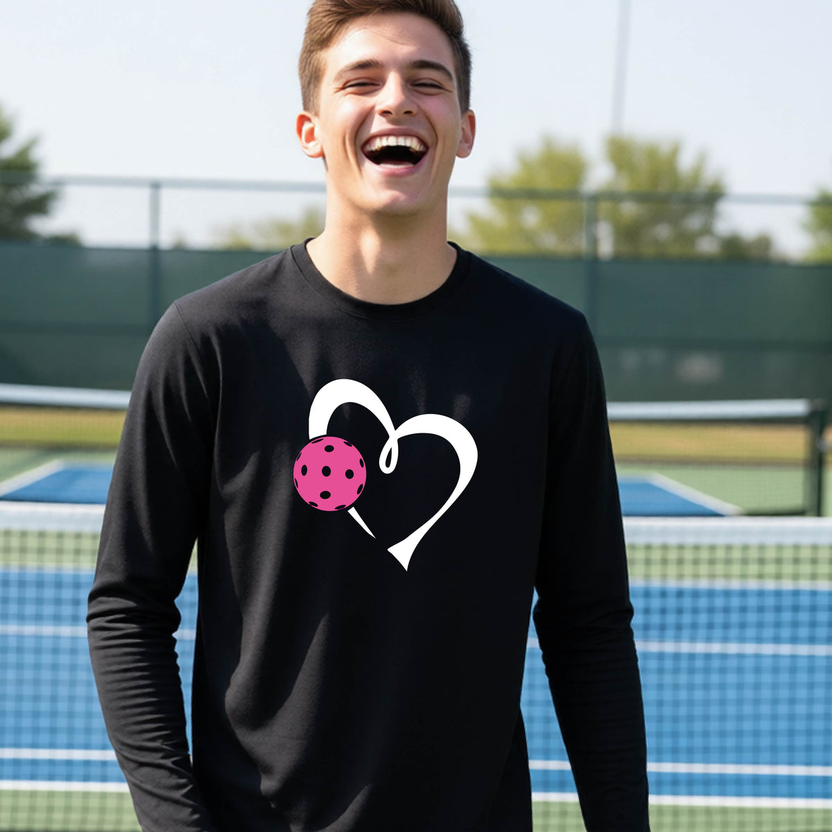A teenager laughing on an outdoor pickleball court, wearing a black long-sleeve shirt with a white heart and pink pickleball graphic,.