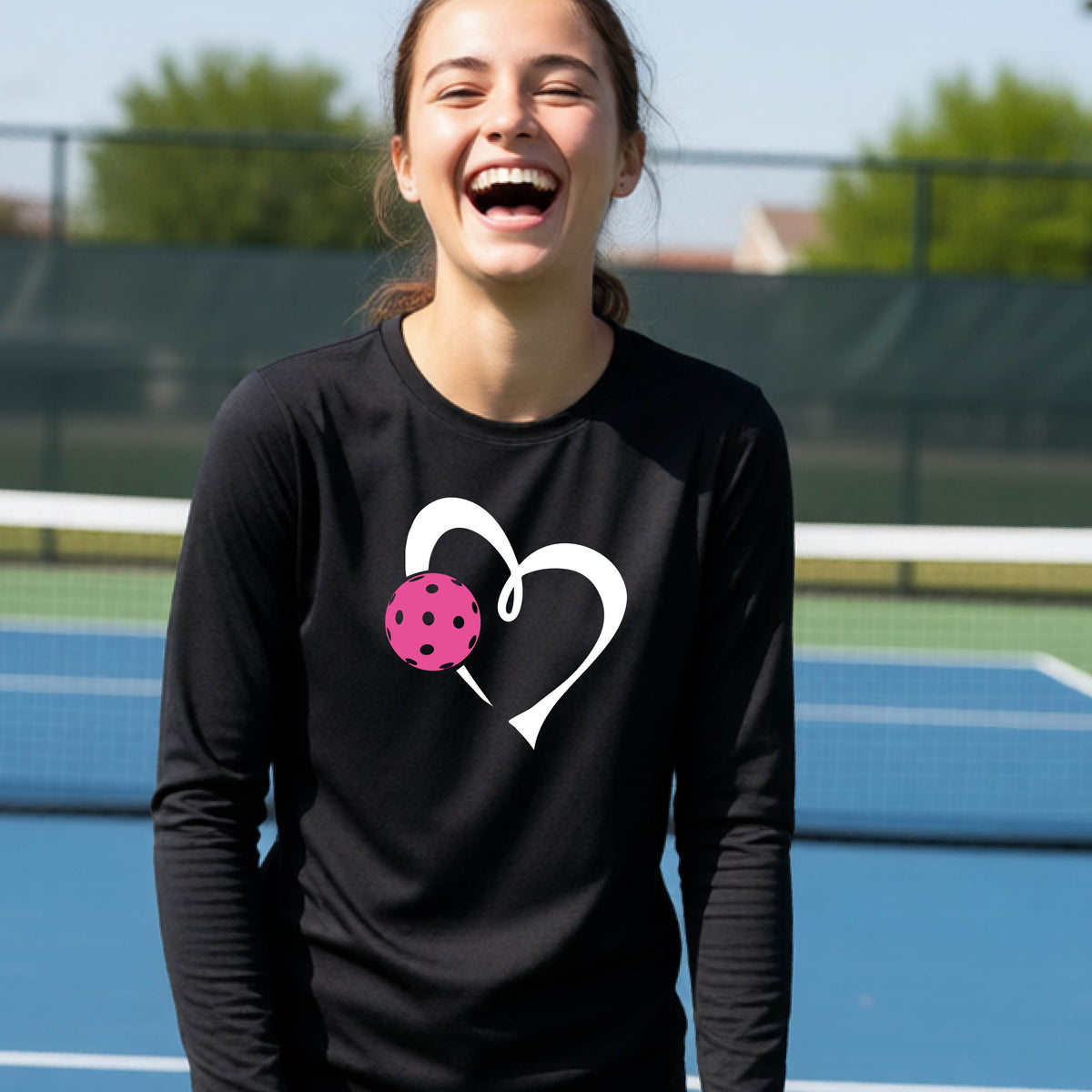 A teenager laughing on an outdoor pickleball court, wearing a black long-sleeve shirt with a white heart and pink pickleball graphic,.