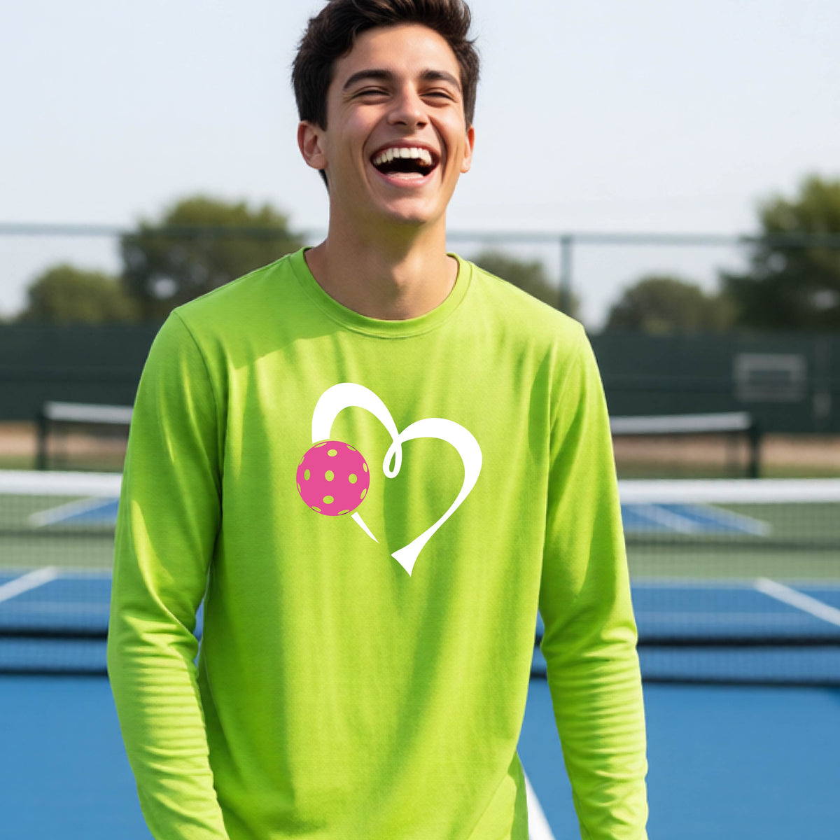 A teenager laughing on an outdoor pickleball court, wearing a lime green long-sleeve shirt with a white heart and pink pickleball graphic,.