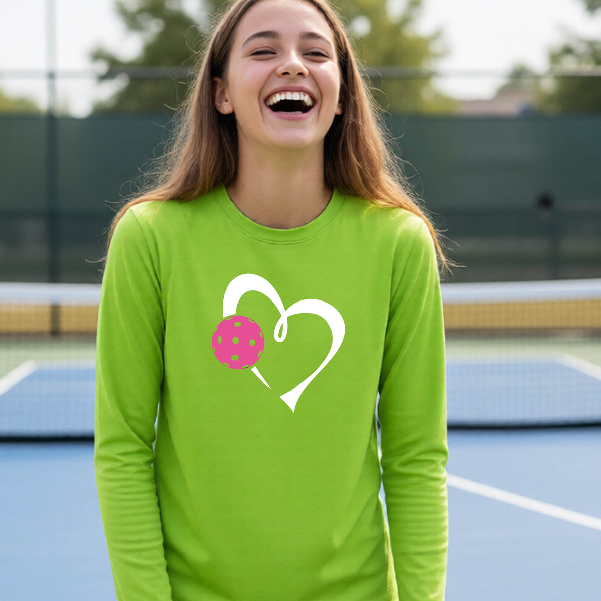 A teenager laughing on an outdoor pickleball court, wearing a lime green long-sleeve shirt with a white heart and pink pickleball graphic,.