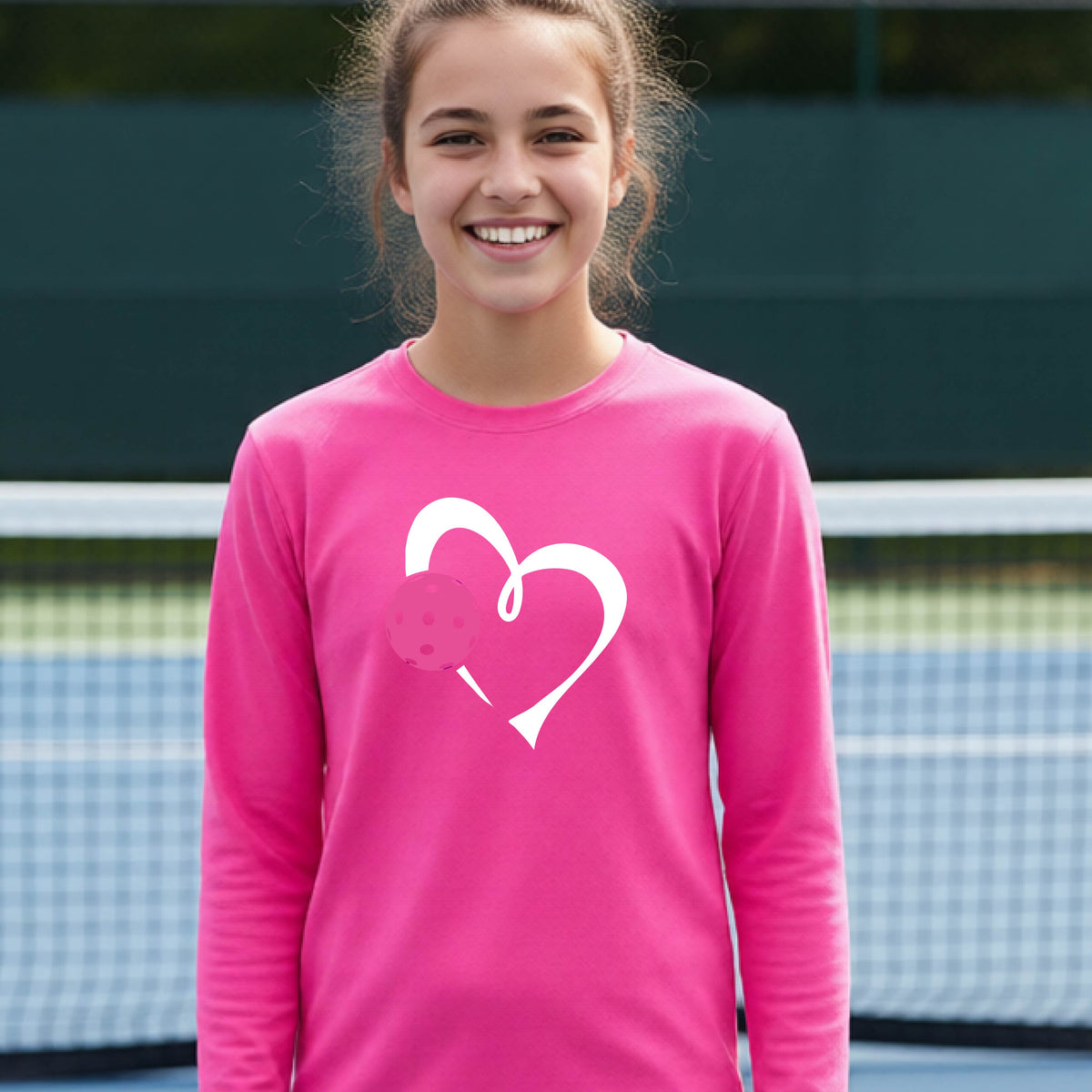 A teenager laughing on an outdoor pickleball court, wearing a pink long-sleeve shirt with a white heart and pink pickleball graphic,.