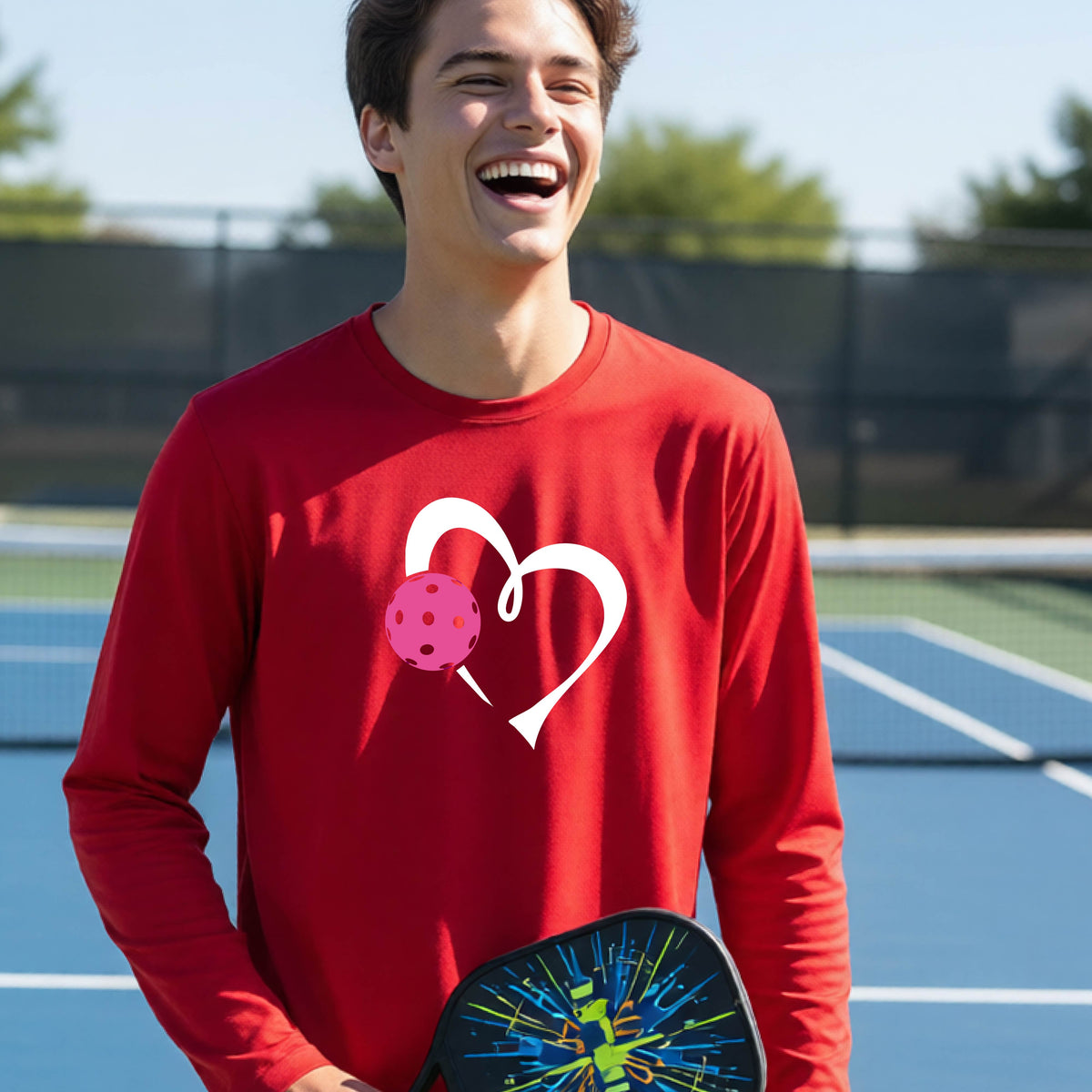 A teenager laughing on an outdoor pickleball court, wearing a red long-sleeve shirt with a white heart and pink pickleball graphic,.