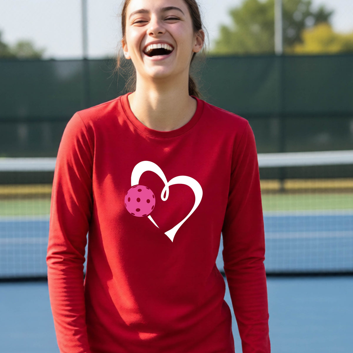 A teenager laughing on an outdoor pickleball court, wearing a red long-sleeve shirt with a white heart and pink pickleball graphic,.