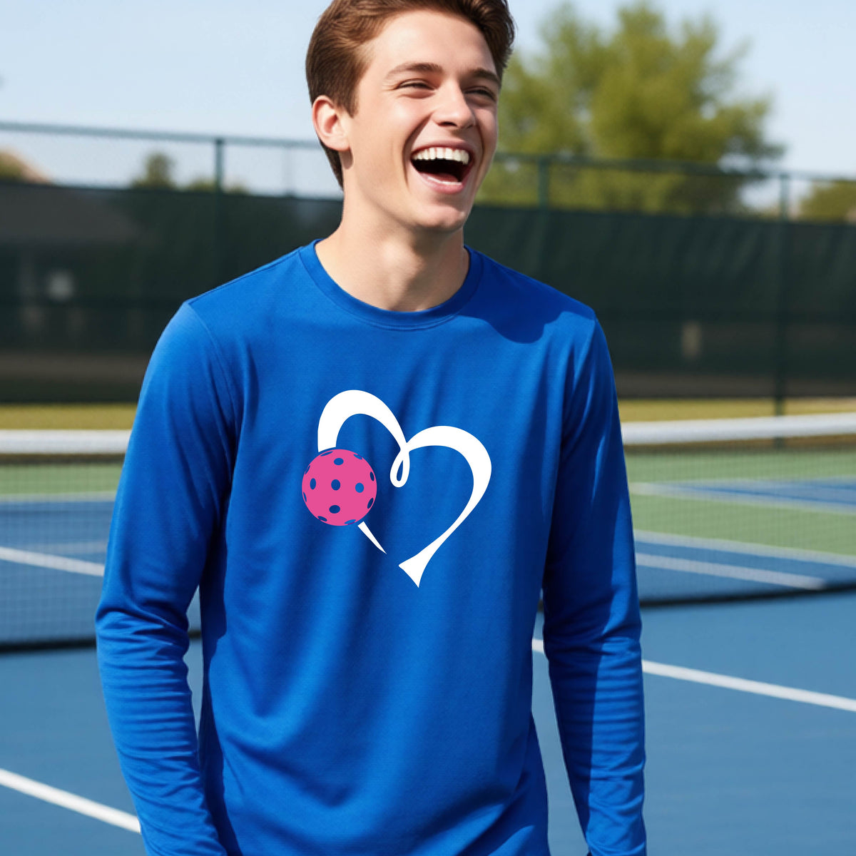 A teenager laughing on an outdoor pickleball court, wearing a royal blue long-sleeve shirt with a white heart and pink pickleball graphic,.