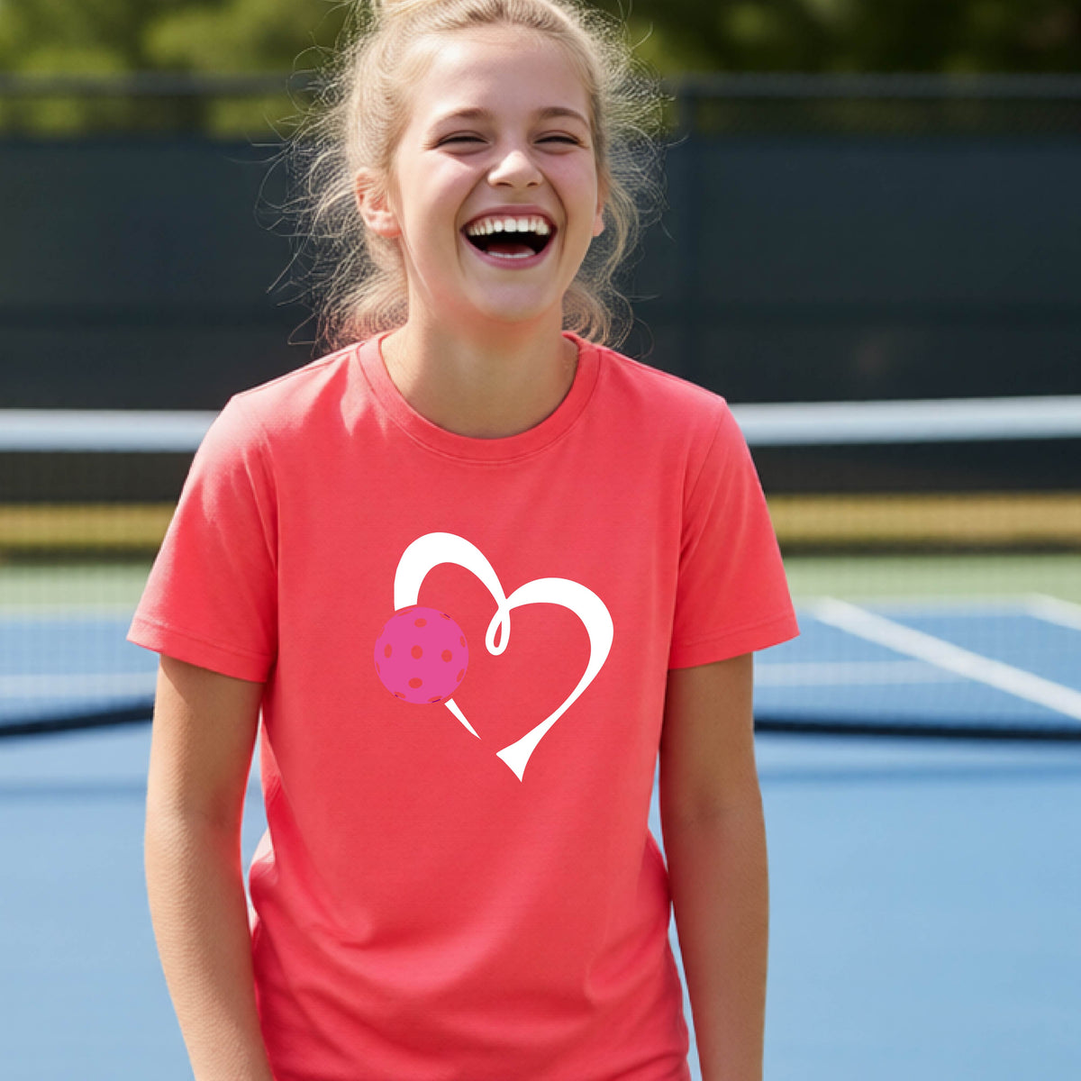 A teenager laughing on an outdoor pickleball court wearing a coral pink t-shirt with a white heart and pink pickleball graphic on the front.