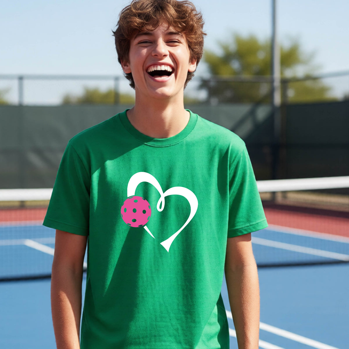 A teenager laughing on an outdoor pickleball court wearing a green t-shirt with a white heart and pink pickleball graphic on the front.