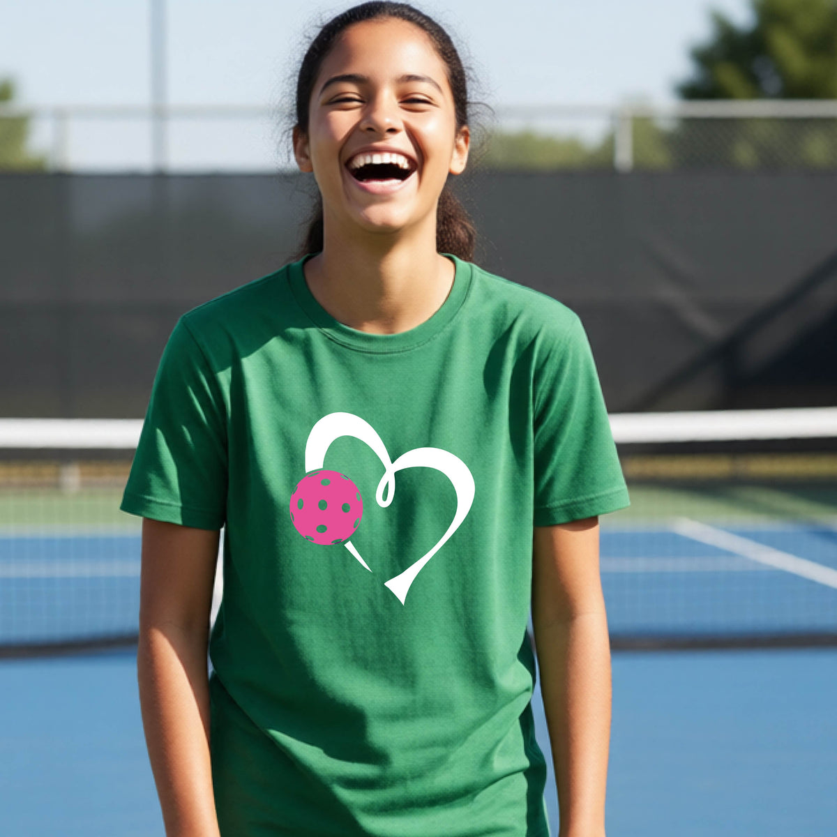 A teenager laughing on an outdoor pickleball court wearing a green t-shirt with a white heart and pink pickleball graphic on the front.