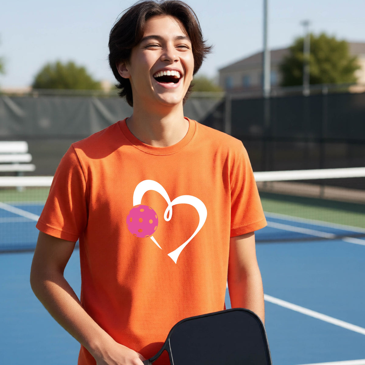 A teenager laughing on an outdoor pickleball court wearing an orange t-shirt with a white heart and pink pickleball graphic on the front.