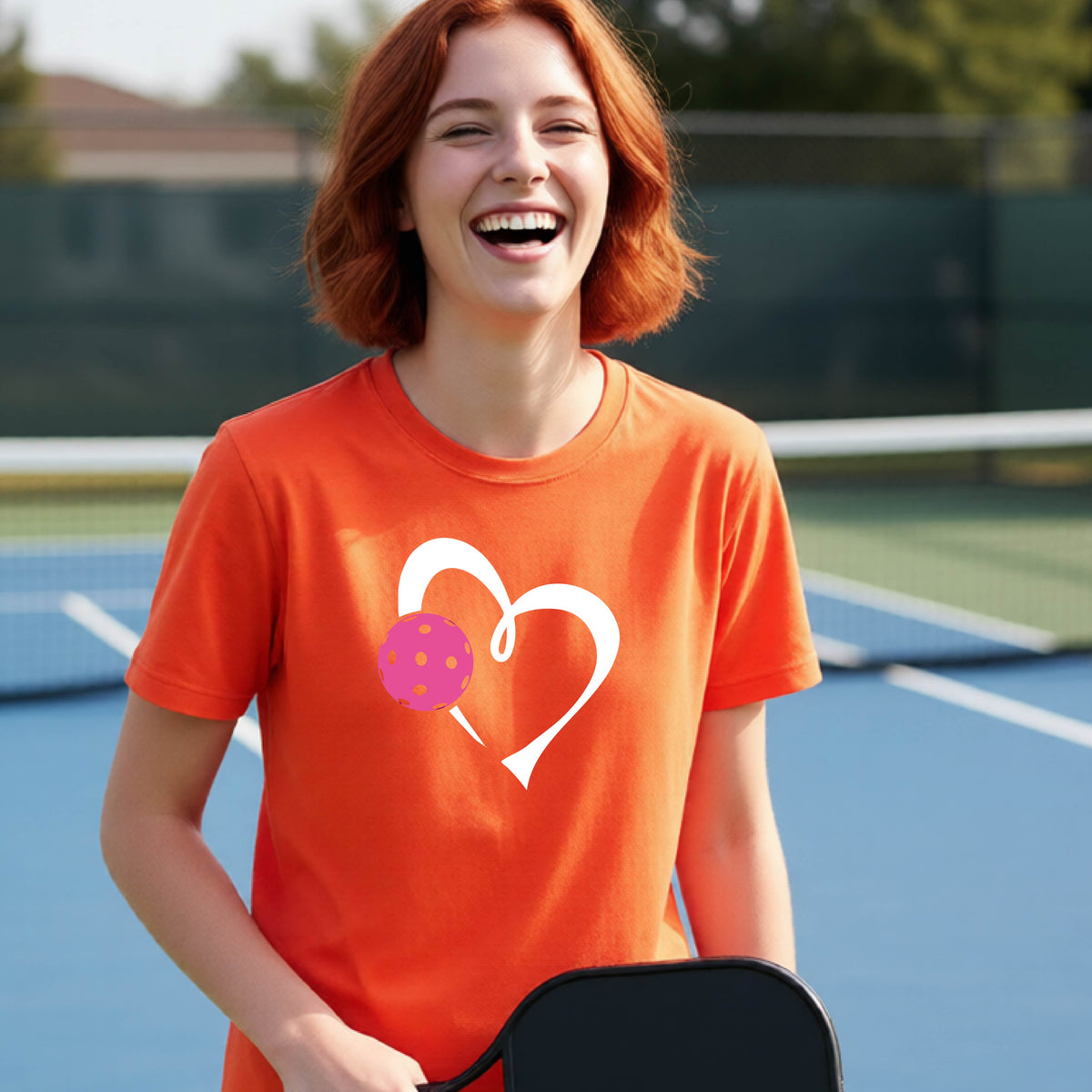 A teenager laughing on an outdoor pickleball court wearing an orange t-shirt with a white heart and pink pickleball graphic on the front.