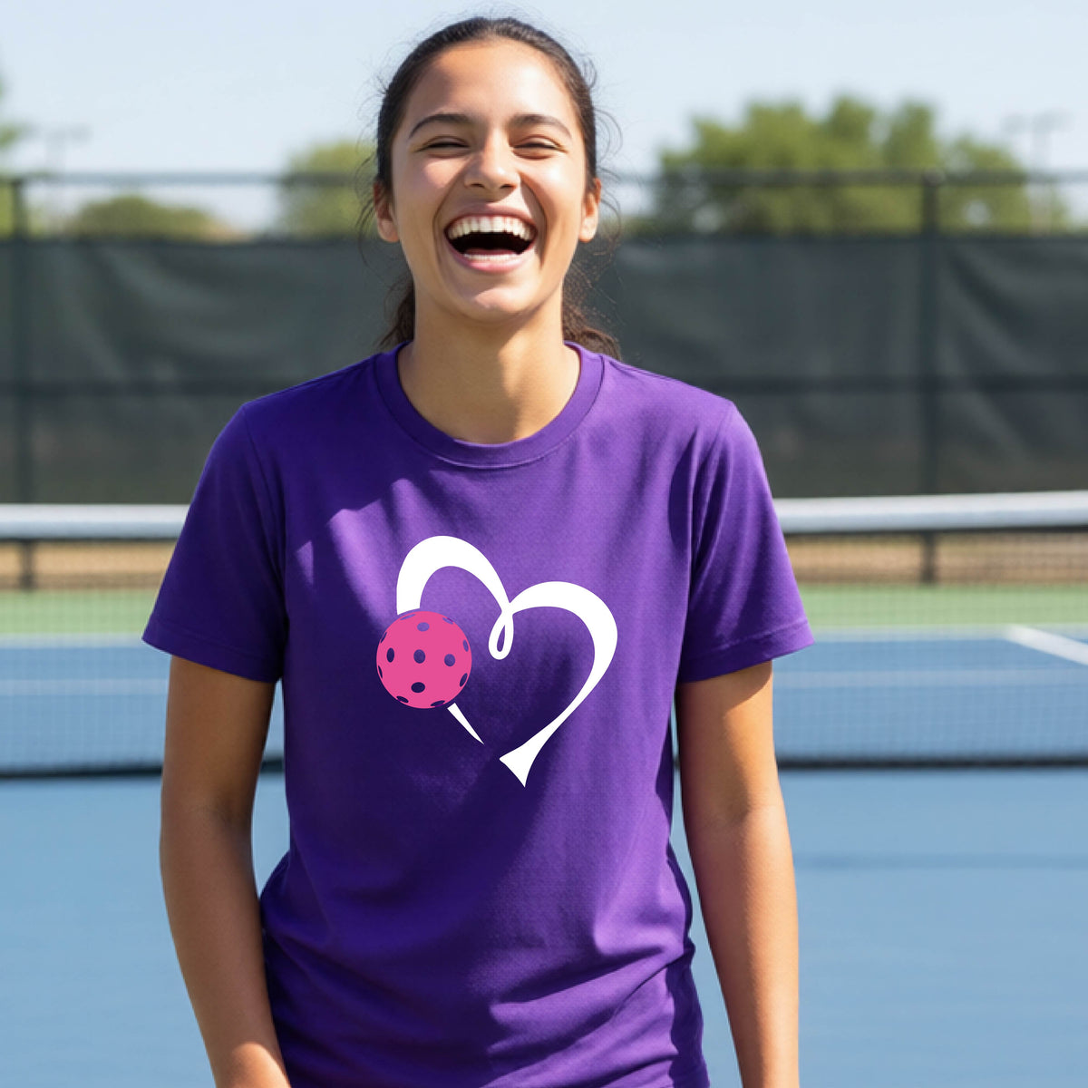 A teenager laughing on an outdoor pickleball court wearing a purple t-shirt with a white heart and pink pickleball graphic on the front.