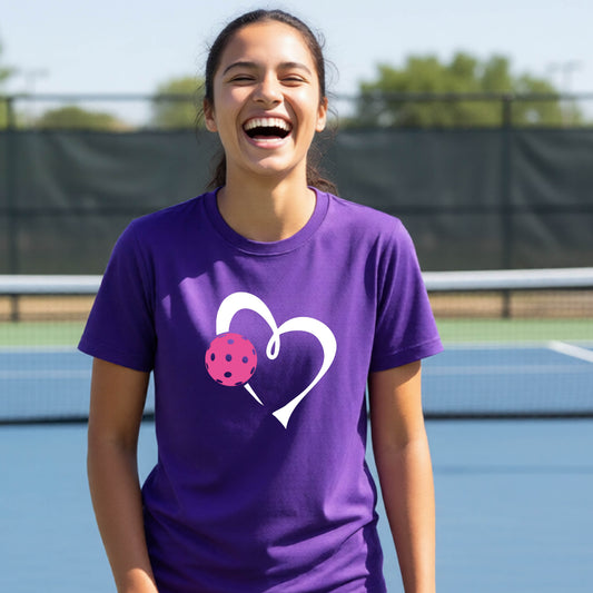 A teenager laughing on an outdoor pickleball court wearing a purple t-shirt with a white heart and pink pickleball graphic on the front.
