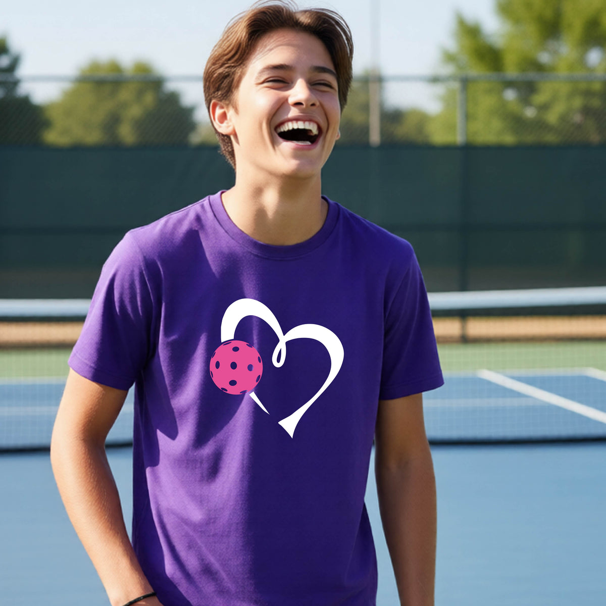 A teenager laughing on an outdoor pickleball court wearing a purple t-shirt with a white heart and pink pickleball graphic on the front.