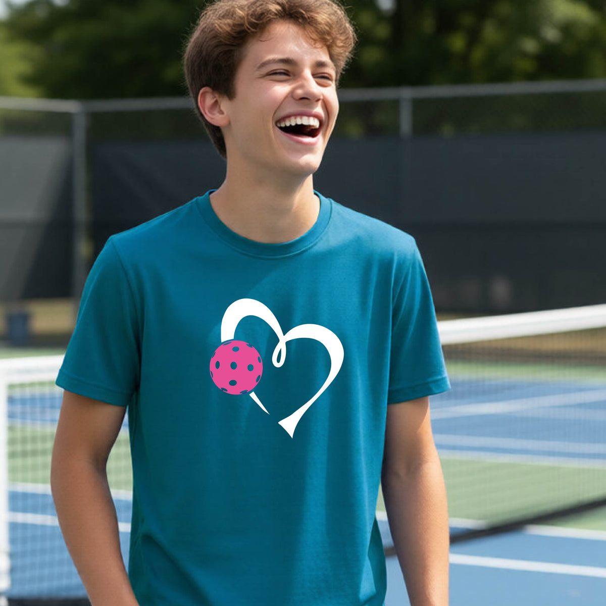 A teenager laughing on an outdoor pickleball court wearing a teal blue t-shirt with a white heart and pink pickleball graphic on the front.