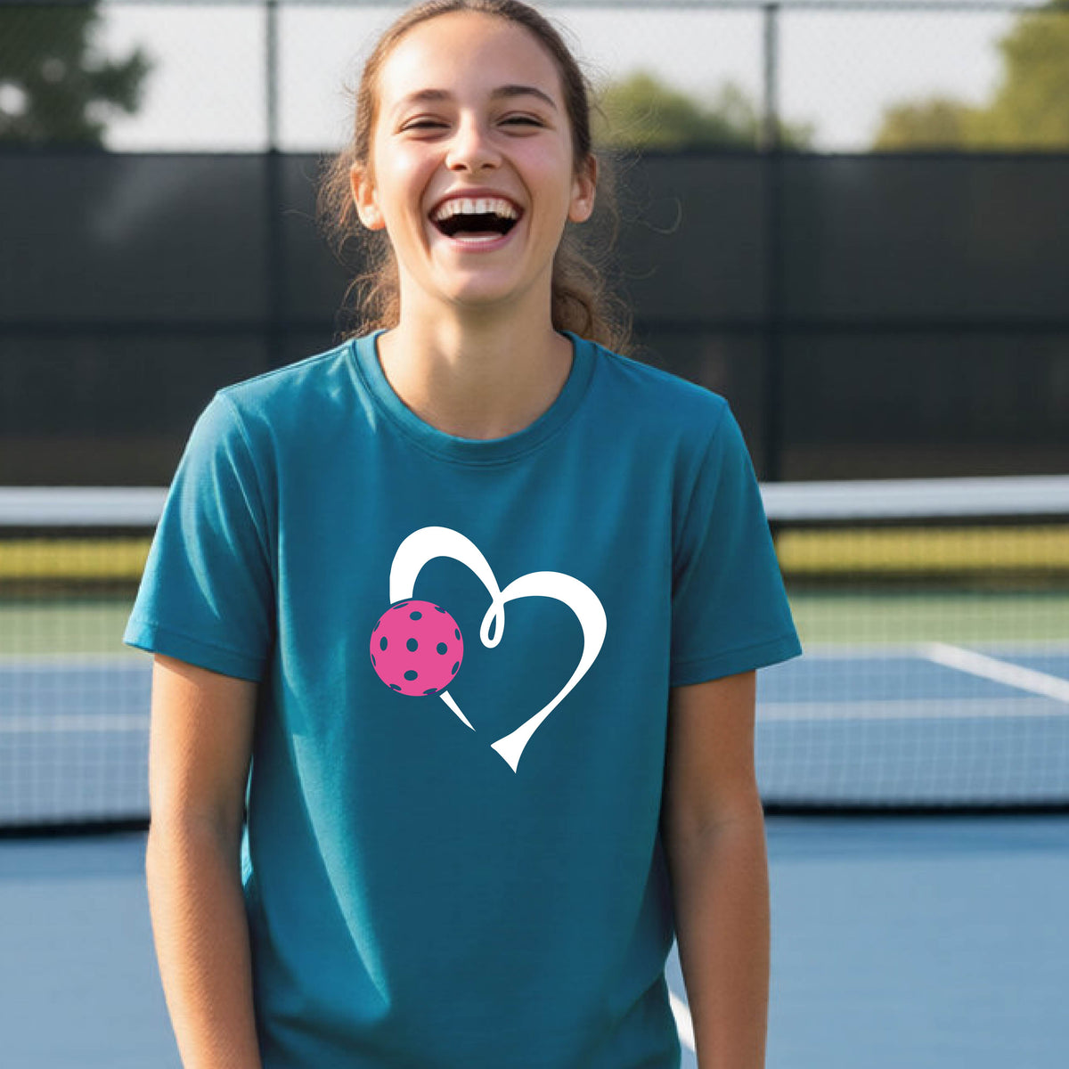 A teenager laughing on an outdoor pickleball court wearing a teal blue t-shirt with a white heart and pink pickleball graphic on the front.
