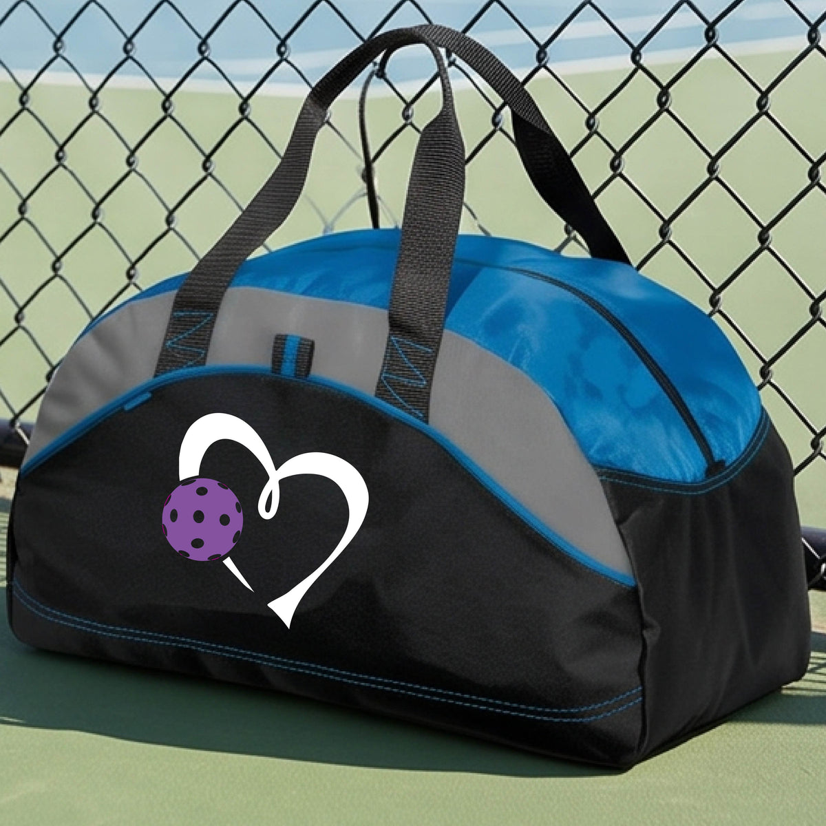 A black and royal blue  pickleball duffel bag with gray panels, featuring a white heart outline and a purple pickleball graphic on the front,.