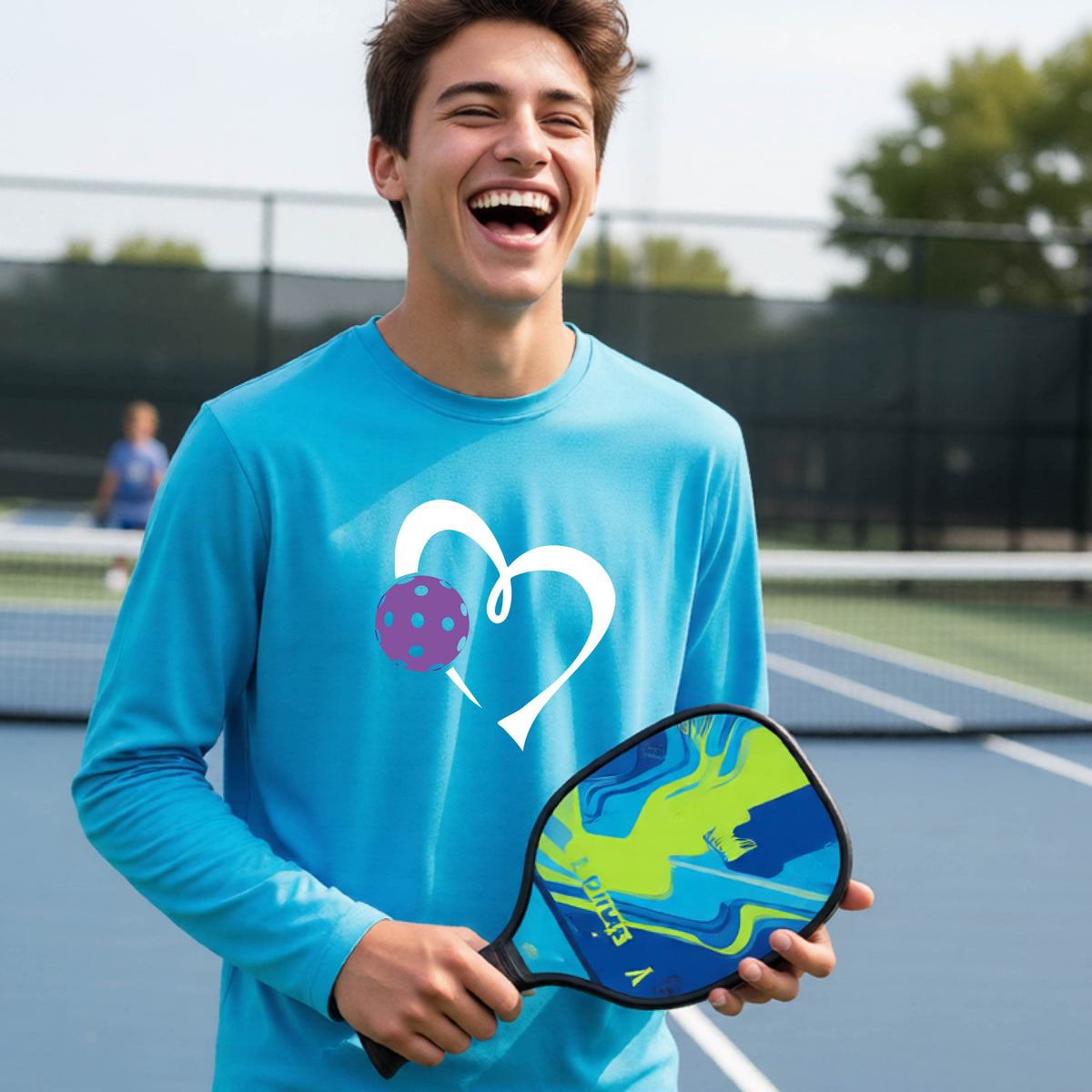 A teenager laughing on a pickelball court wearing a bright blue long-sleeve shirt with a white heart outline and a purple pickleball graphic.