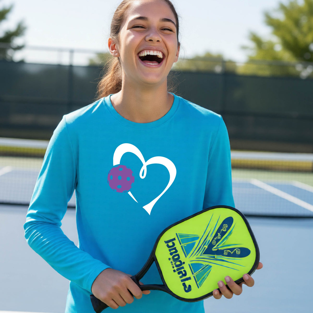 A teenager laughing on a pickelball court wearing a bright blue long-sleeve shirt with a white heart outline and a purple pickleball graphic.