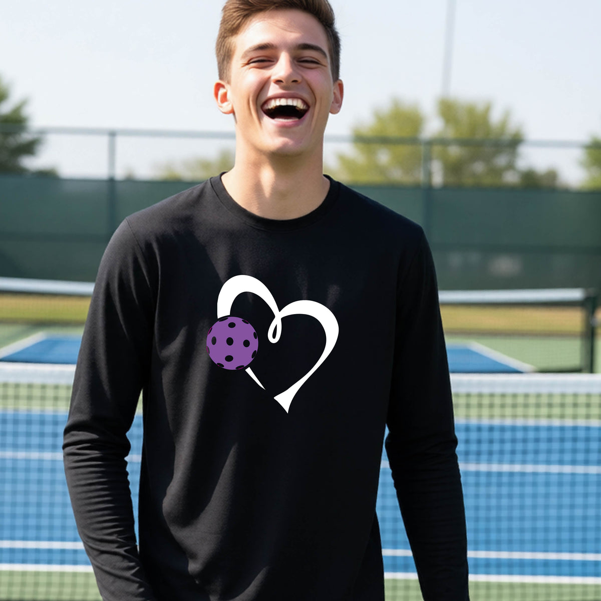 A teenager laughing on a pickelball court wearing a black long-sleeve shirt with a white heart outline and a purple pickleball graphic.