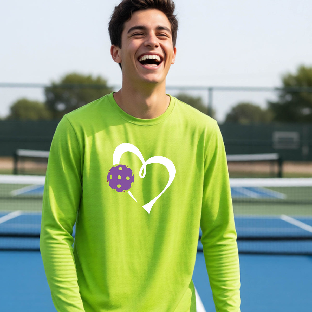 A teenager laughing on a pickelball court wearing a lime green long-sleeve shirt with a white heart outline and a purple pickleball graphic.