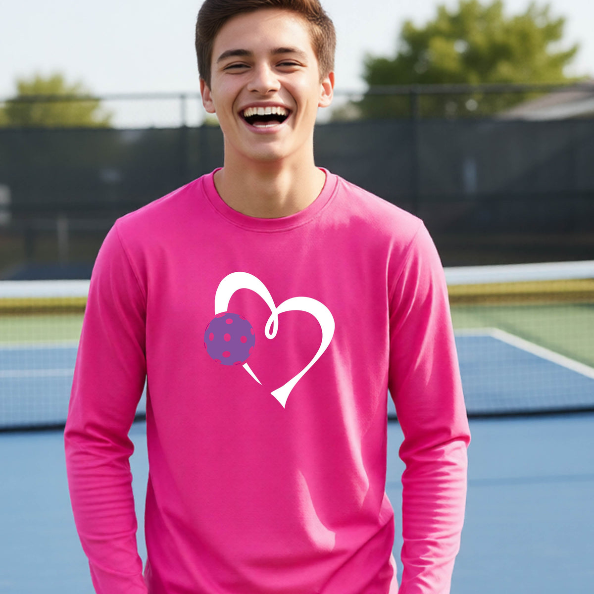 A teenager laughing on a pickelball court wearing a pink long-sleeve shirt with a white heart outline and a purple pickleball graphic.