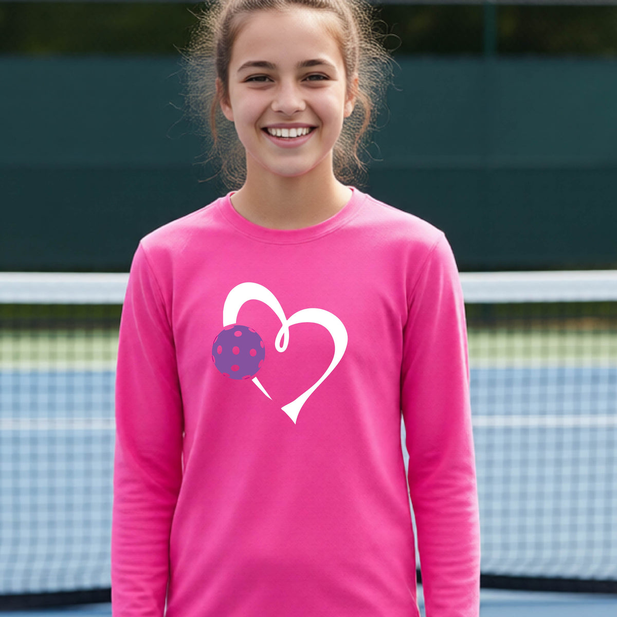 A teenager laughing on a pickelball court wearing a pink long-sleeve shirt with a white heart outline and a purple pickleball graphic.