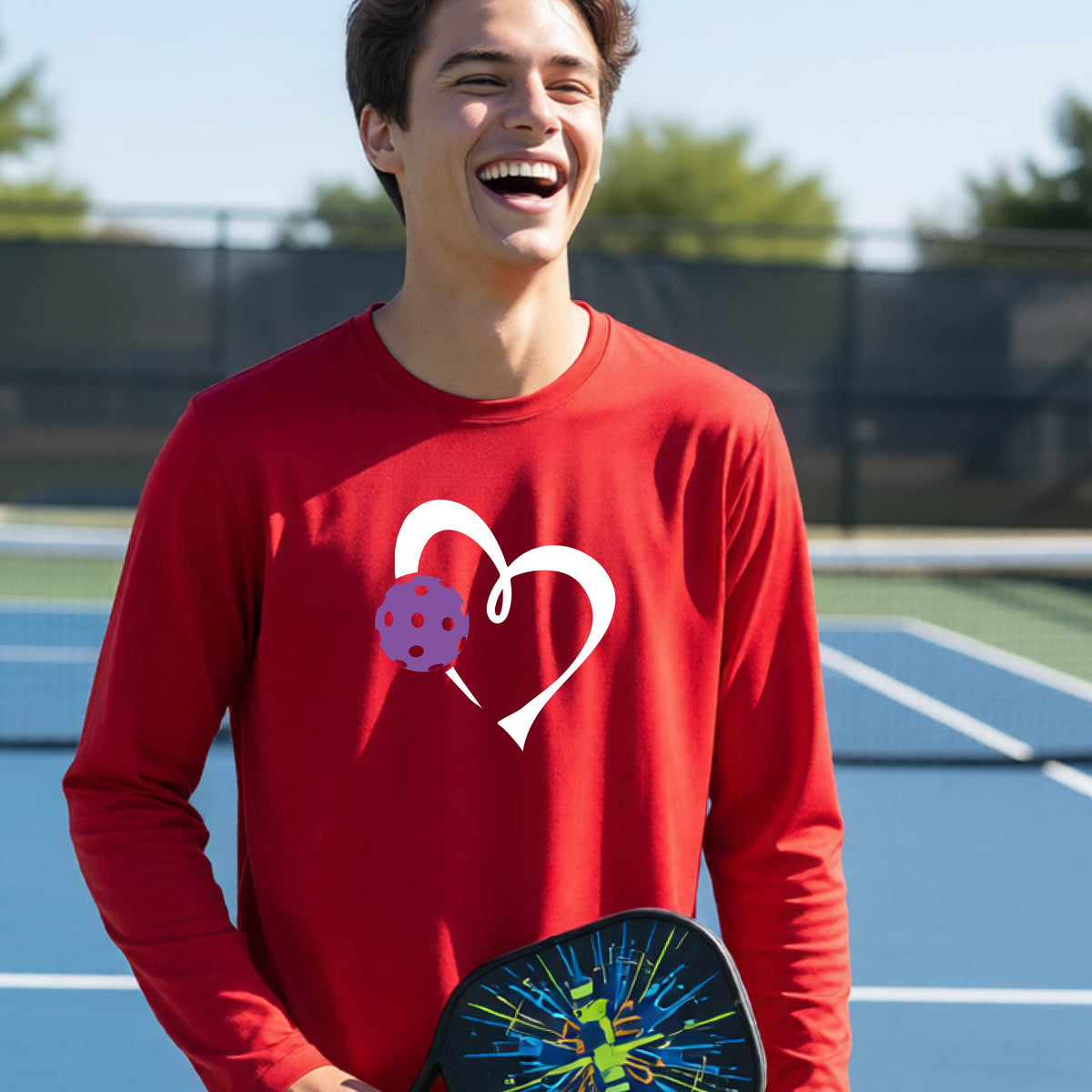A teenager laughing on a pickelball court wearing a red long-sleeve shirt with a white heart outline and a purple pickleball graphic.