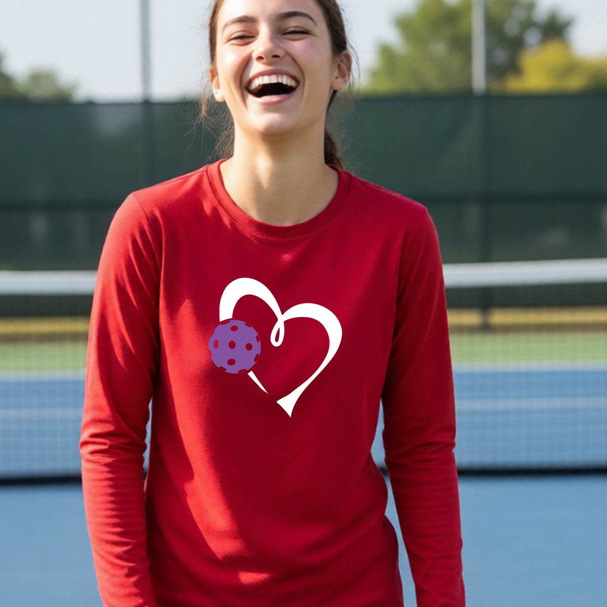 A teenager laughing on a pickelball court wearing a red long-sleeve shirt with a white heart outline and a purple pickleball graphic.