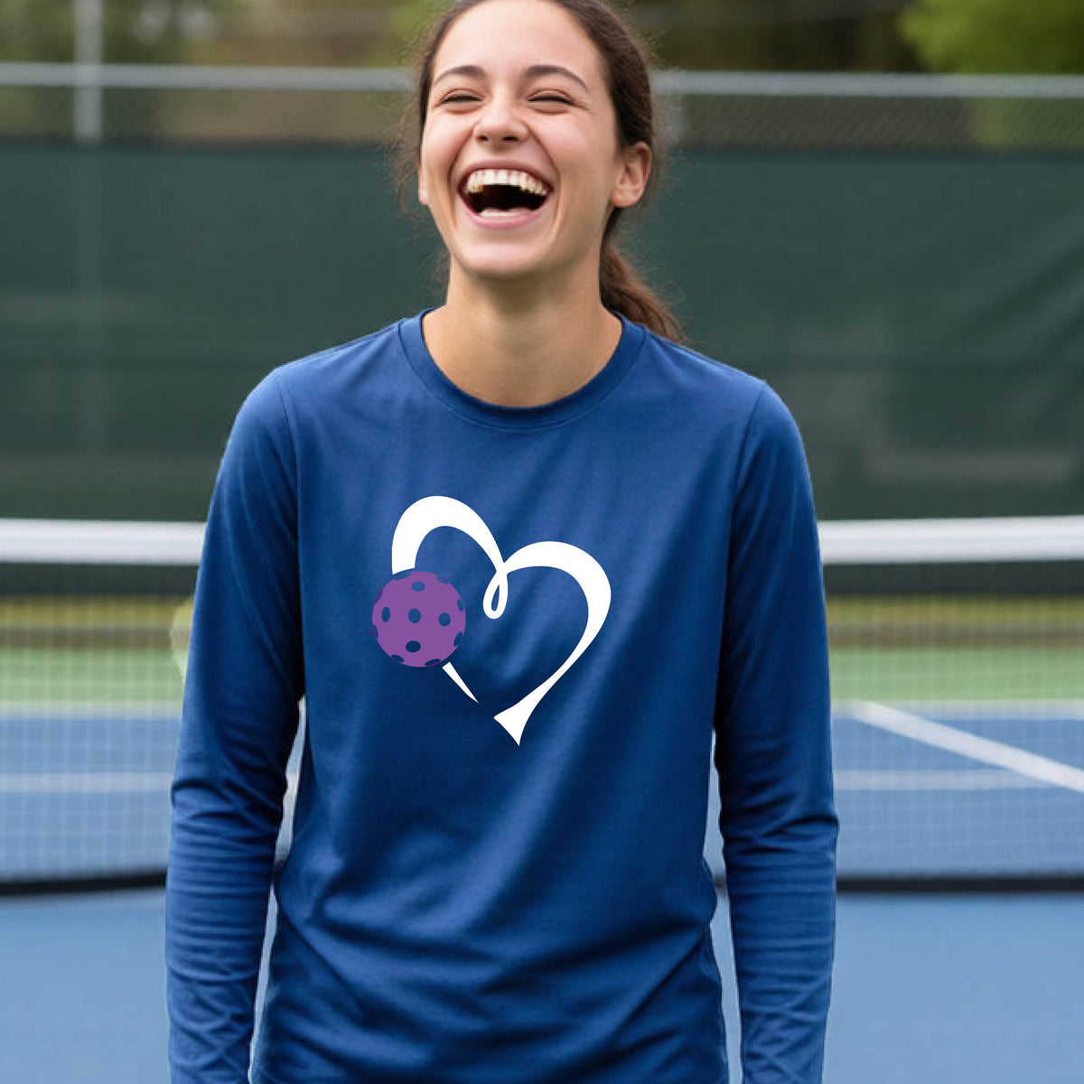 A teenager laughing on a pickelball court wearing a royal blue long-sleeve shirt with a white heart outline and a purple pickleball graphic.