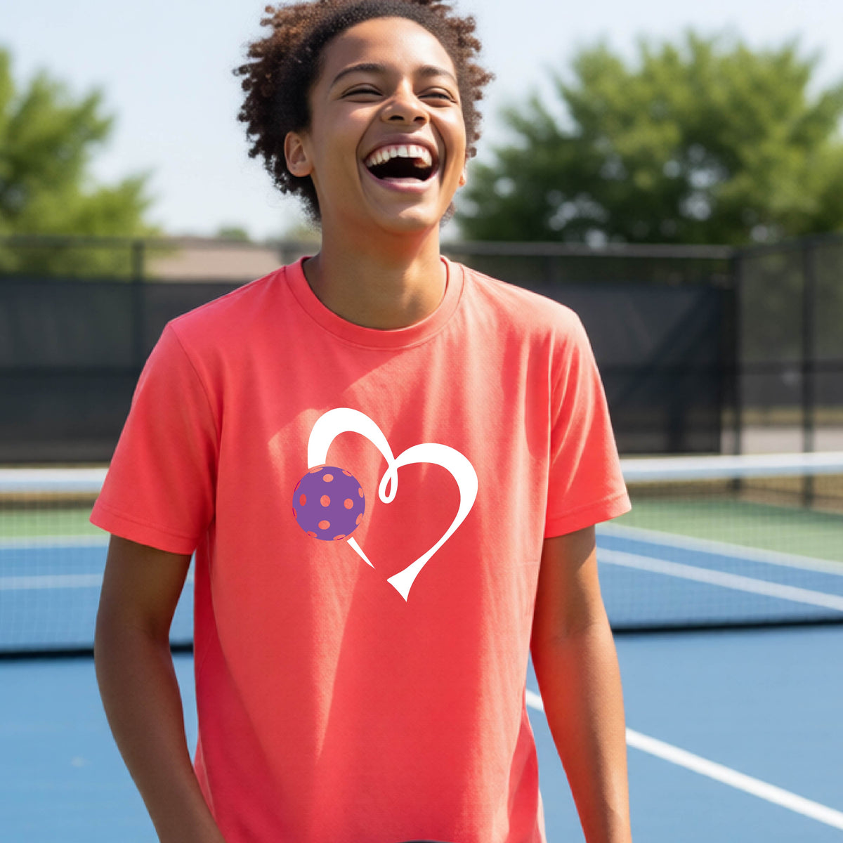 A young teenager laughing on a pickelball court, wearing a coral pink t-shirt with a white heart outline and a purple pickleball graphic.