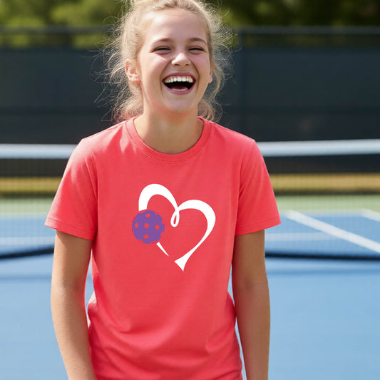 A young teenager laughing on a pickelball court, wearing a coral pink t-shirt with a white heart outline and a purple pickleball graphic.