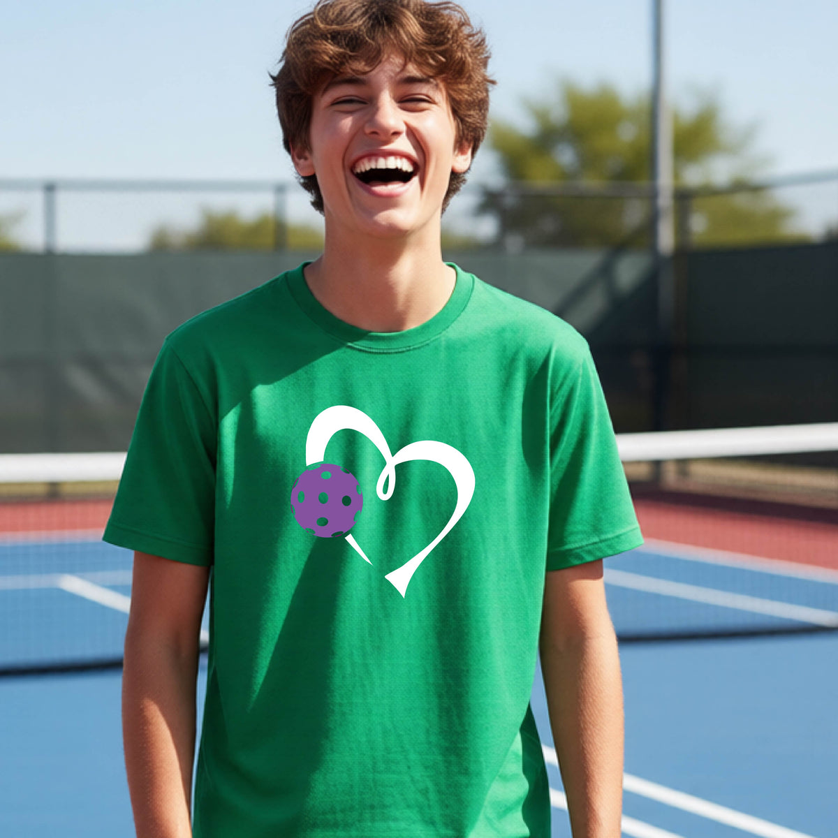 A young teenager laughing on a pickelball court, wearing a green t-shirt with a white heart outline and a purple pickleball graphic.