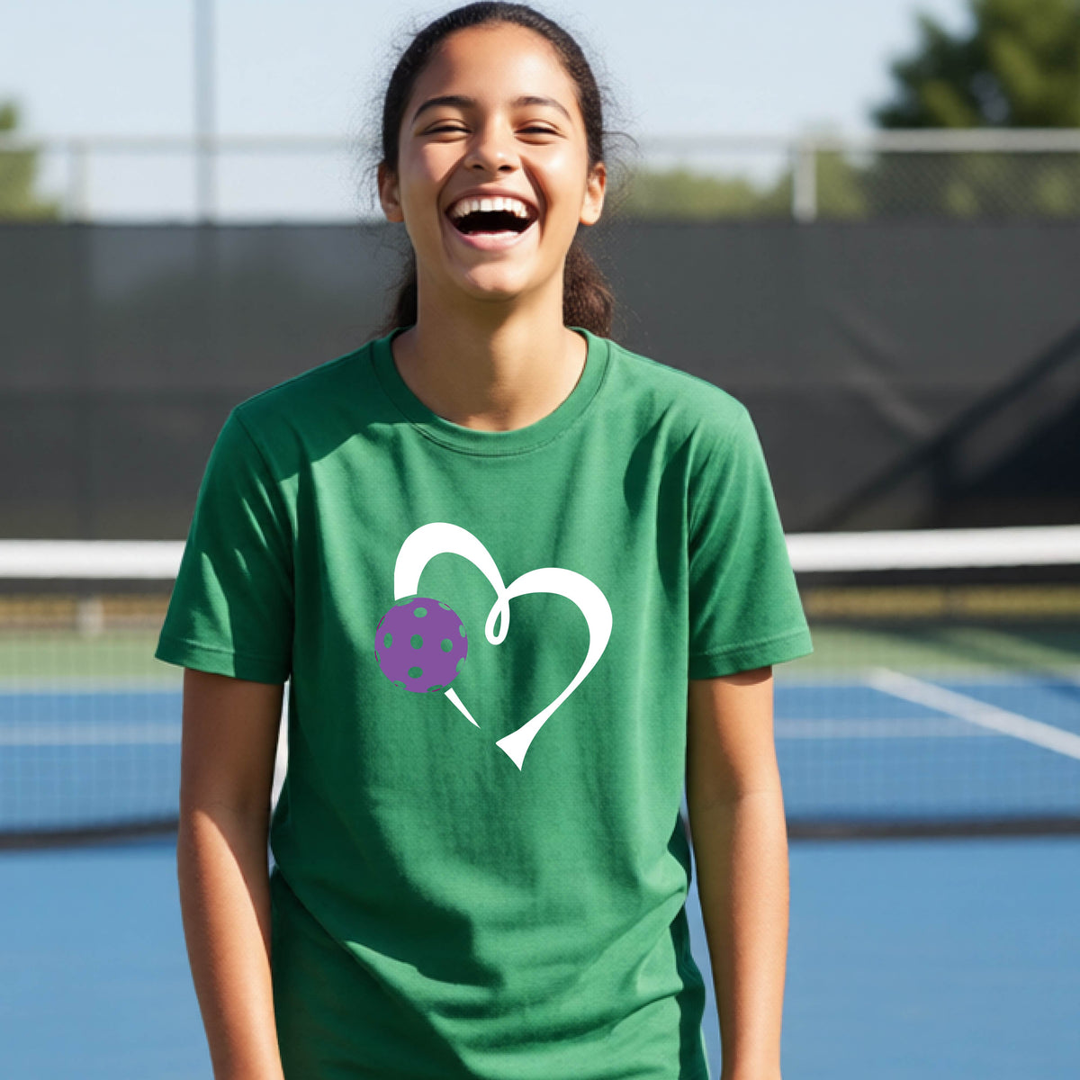A young teenager laughing on a pickelball court, wearing a green t-shirt with a white heart outline and a purple pickleball graphic.