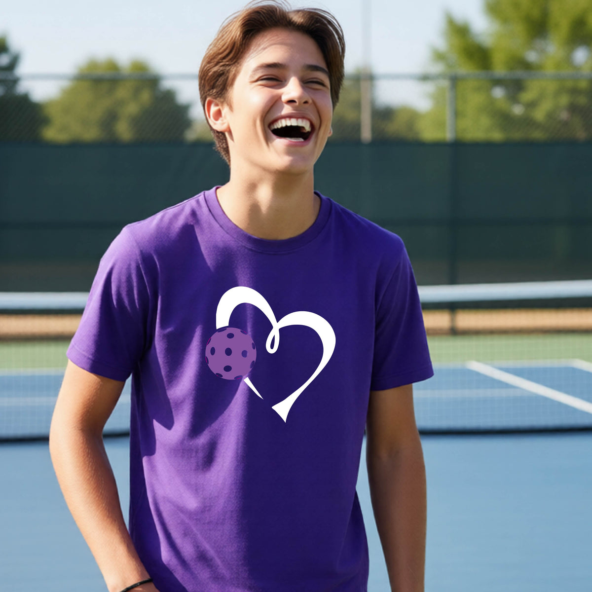 A young teenager laughing on a pickelball court, wearing a purple t-shirt with a white heart outline and a purple pickleball graphic.