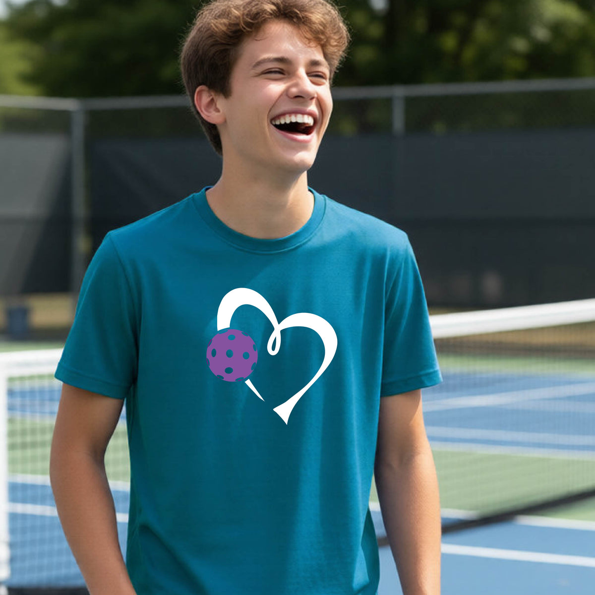 A young teenager laughing on a pickelball court, wearing a teal blue t-shirt with a white heart outline and a purple pickleball graphic.