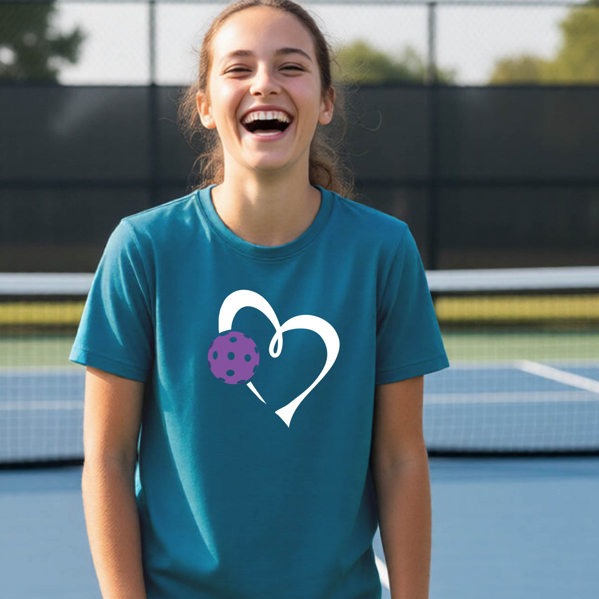 A young teenager laughing on a pickelball court, wearing a teal blue t-shirt with a white heart outline and a purple pickleball graphic.