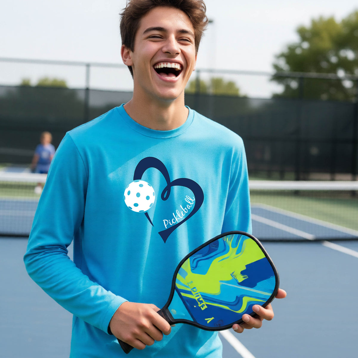 A young person wearing a bright blue long-sleeve athletic t-shirt with a navy blue heart, a white pickleball, and the text 'Pickleball' in script on the front of the shirt. 