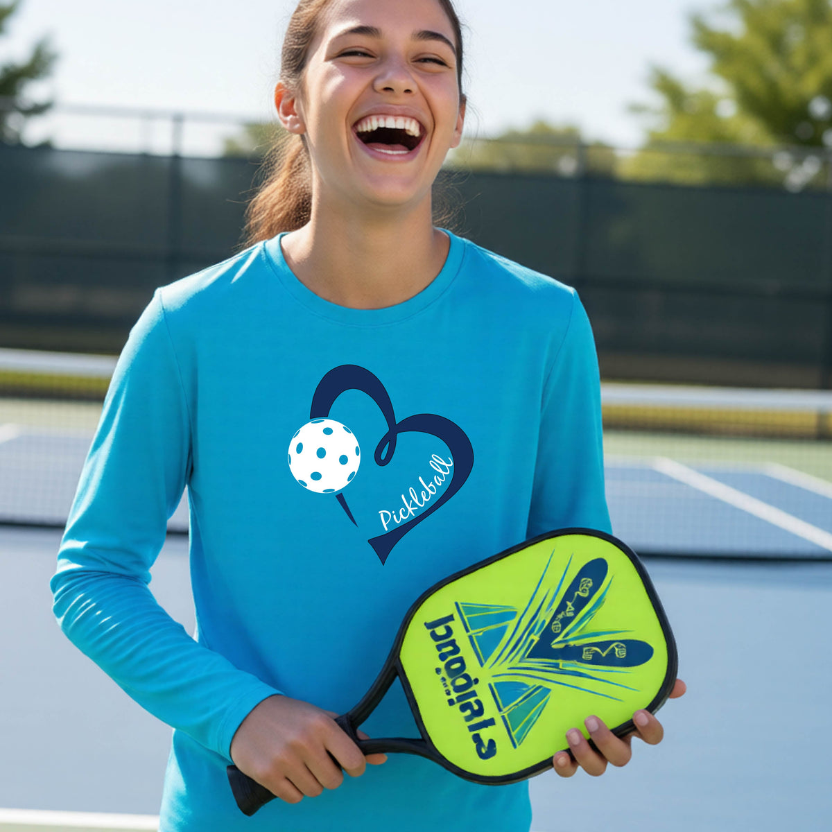 A young person wearing a bright blue long-sleeve athletic t-shirt with a navy blue heart, a white pickleball, and the text 'Pickleball' in script on the front of the shirt. 