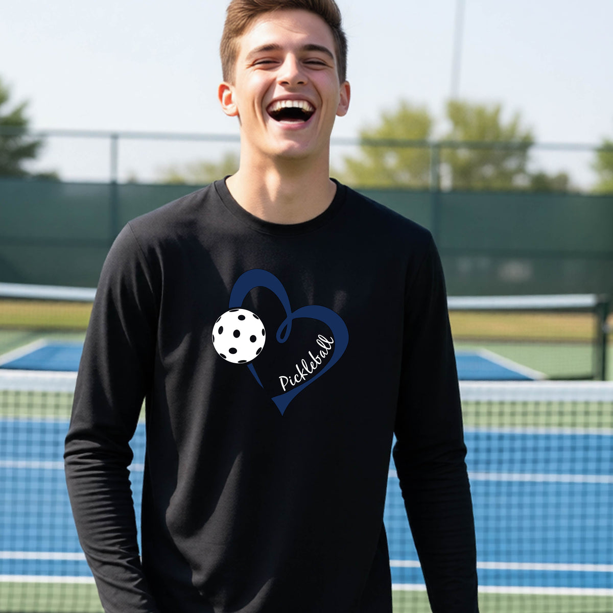A young person wearing a black long-sleeve athletic t-shirt with a navy blue heart, a white pickleball, and the text 'Pickleball' in script on the front of the shirt. 