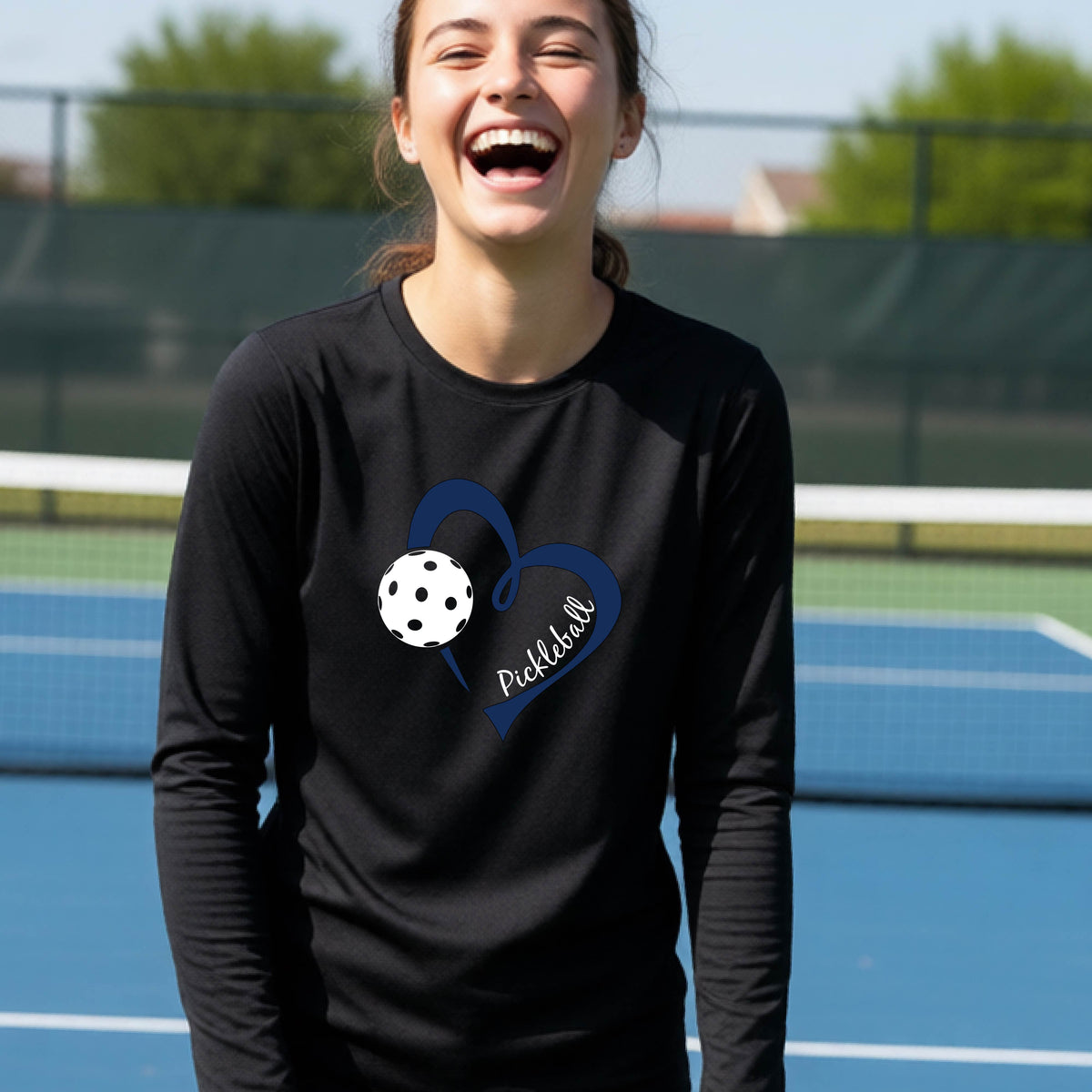 A young person wearing a black long-sleeve athletic t-shirt with a navy blue heart, a white pickleball, and the text 'Pickleball' in script on the front of the shirt. 