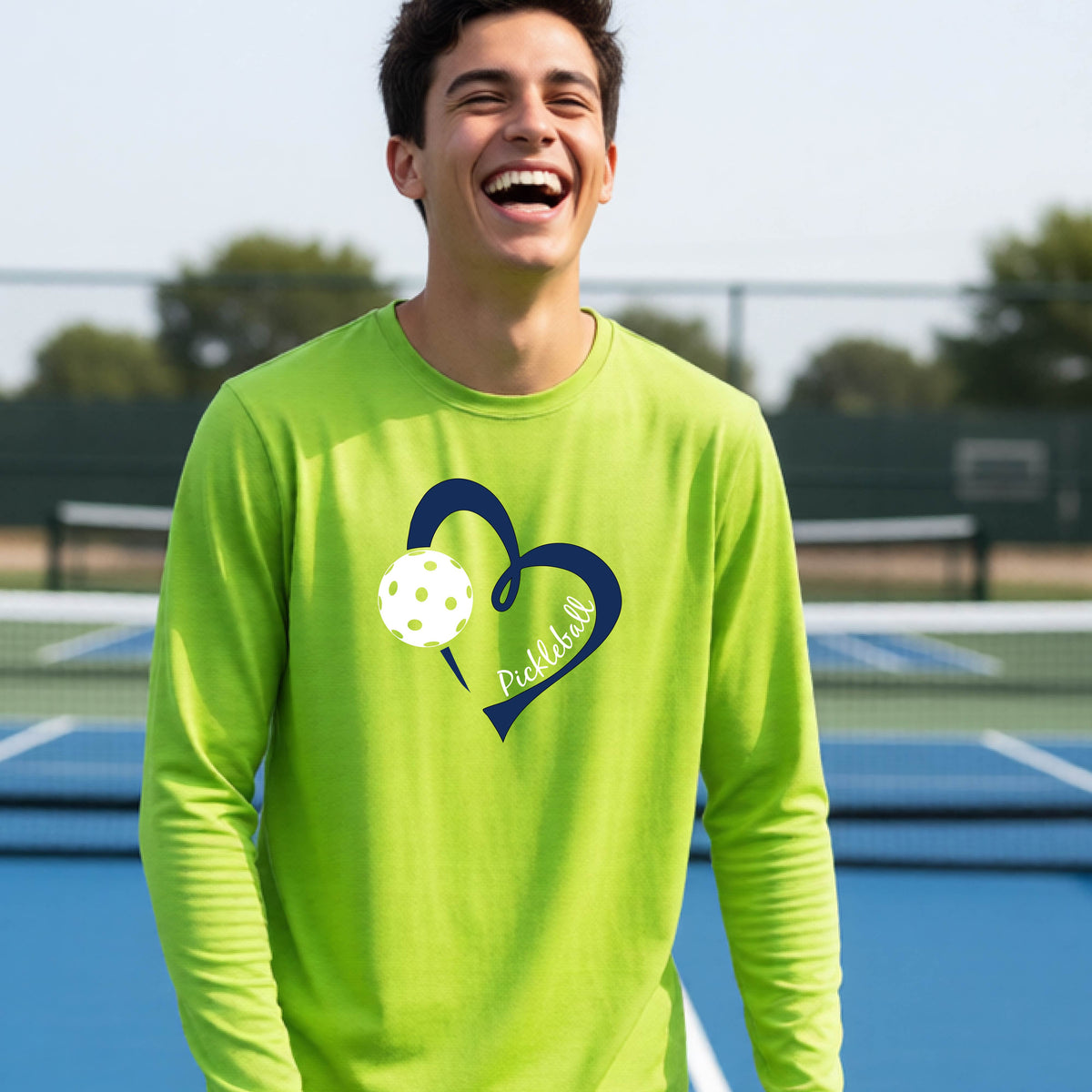 A young person wearing a lime green long-sleeve athletic t-shirt with a navy blue heart, a white pickleball, and the text 'Pickleball' in script on the front of the shirt. 
