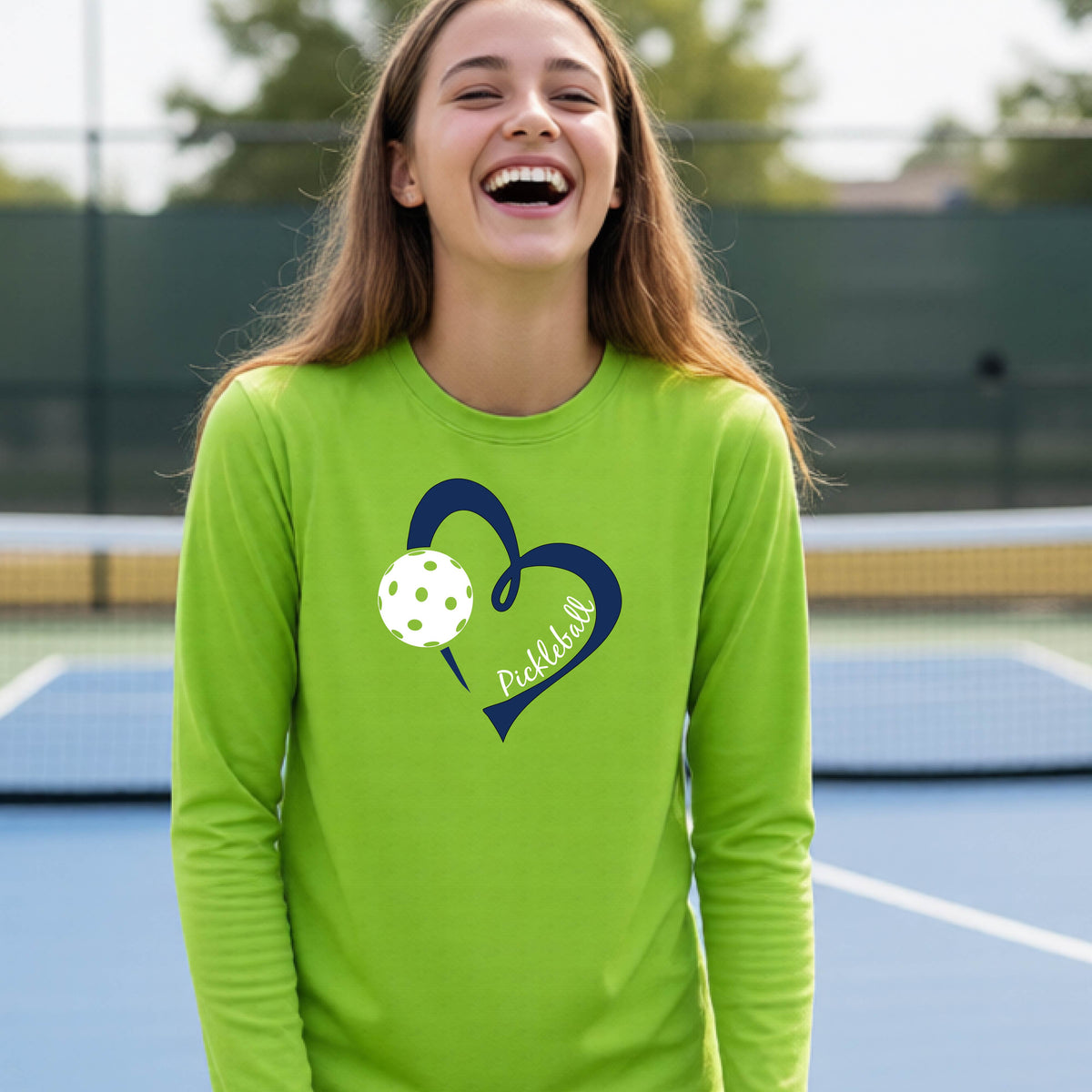 A young person wearing a lime green long-sleeve athletic t-shirt with a navy blue heart, a white pickleball, and the text 'Pickleball' in script on the front of the shirt. 