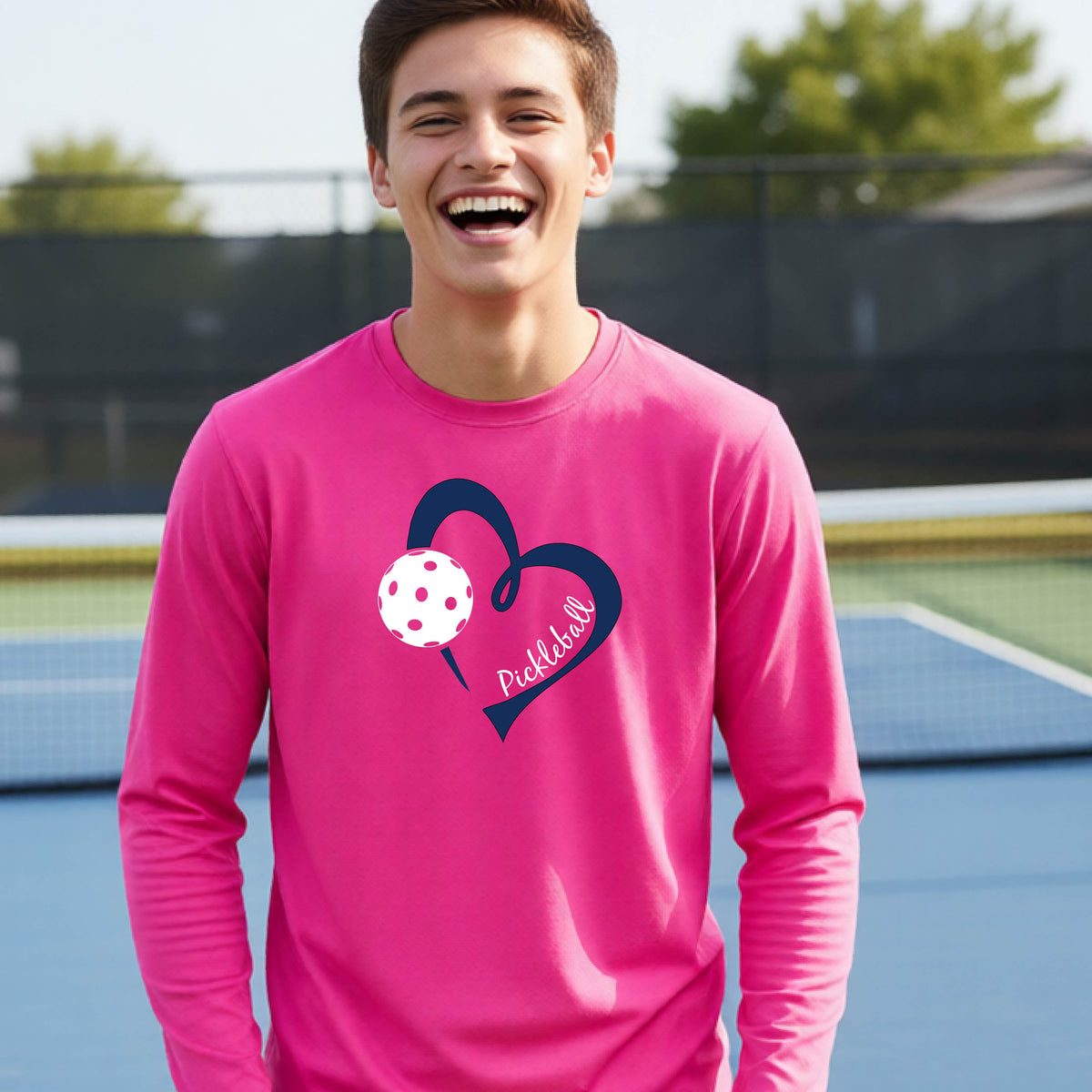 A young person wearing a pink long-sleeve athletic t-shirt with a navy blue heart, a white pickleball, and the text 'Pickleball' in script on the front of the shirt. 