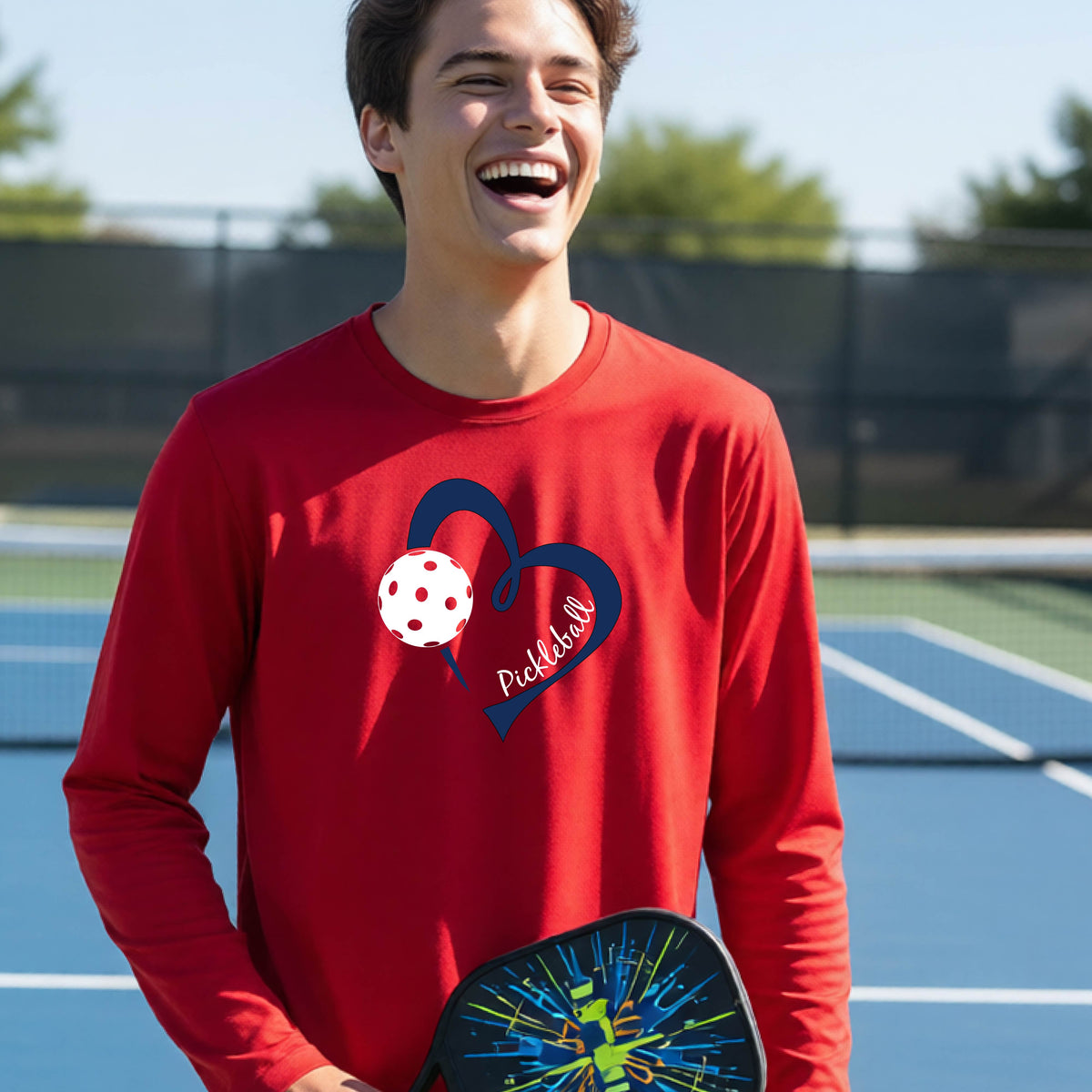 A young person wearing a red long-sleeve athletic t-shirt with a navy blue heart, a white pickleball, and the text 'Pickleball' in script on the front of the shirt. 