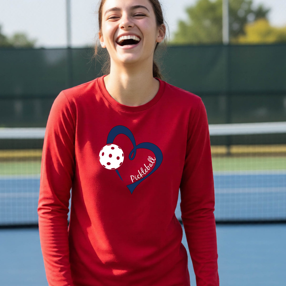 A young person wearing a red long-sleeve athletic t-shirt with a navy blue heart, a white pickleball, and the text 'Pickleball' in script on the front of the shirt. 
